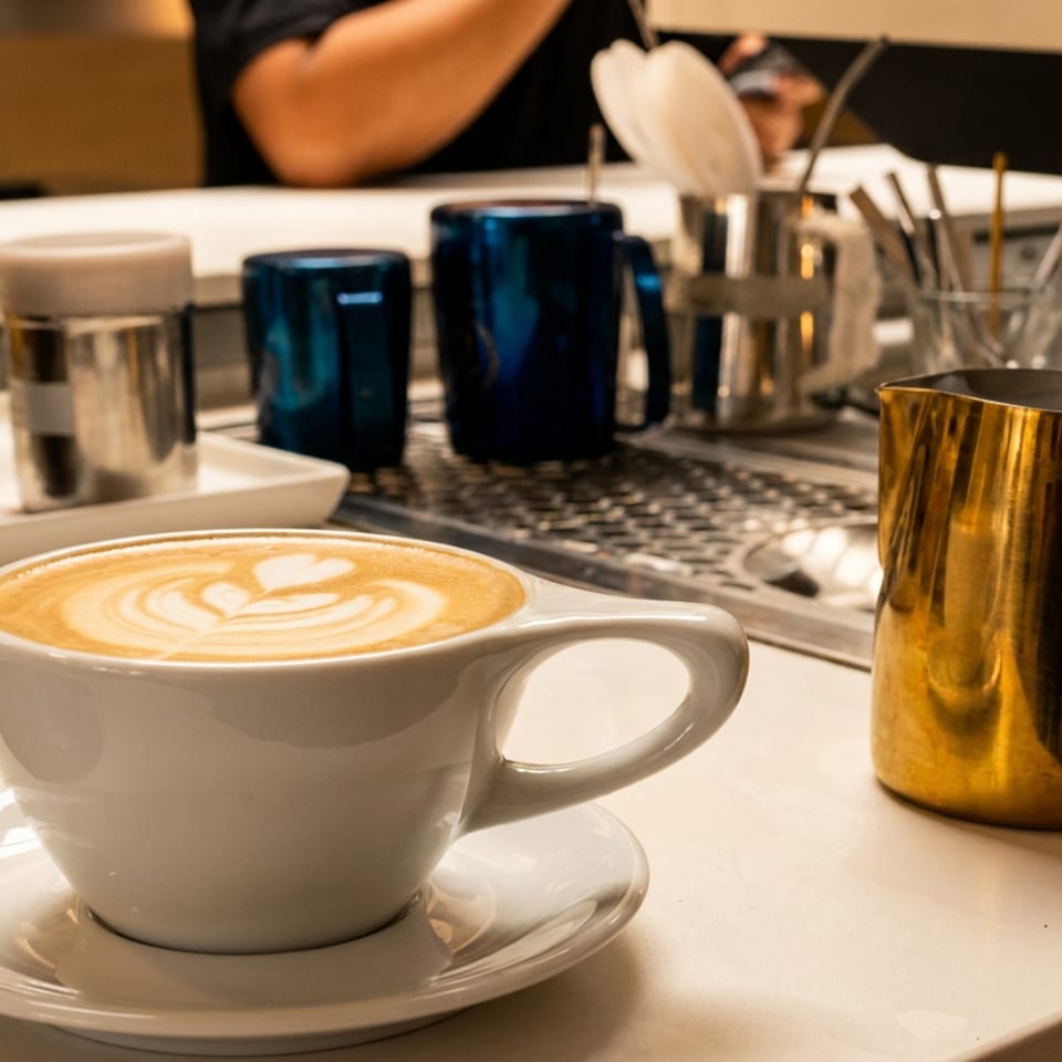 Latte art in a white cup sits on a café counter beside a gold milk pitcher, blue mugs, and coffee supplies.